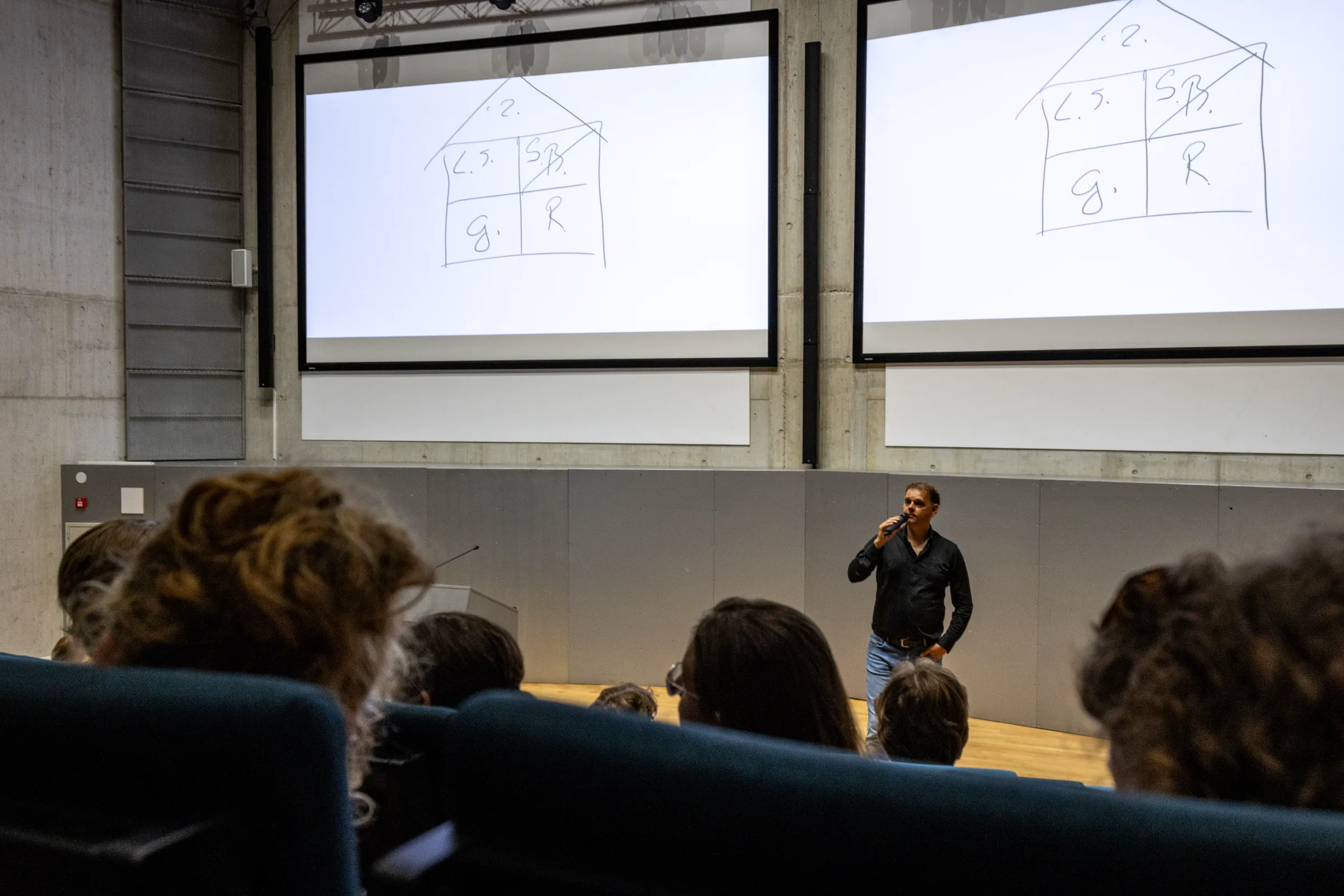 Ronnie Couwenberg staat met een microfoon voor een zaal, op de schermen achter hem staat een getekend huisje.
